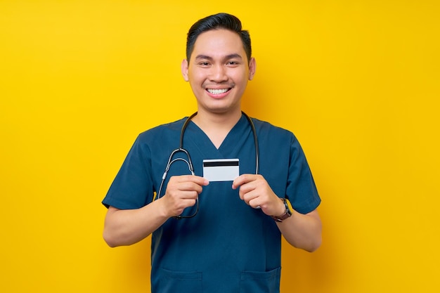 Portrait of a smiling cheerful young asian male doctor or nurse wearing blue uniform holding a credit bank card isolated on yellow background healthcare medicine concept