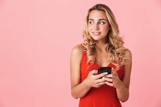 Portrait of a smiling cheery happy emotional cute young pretty blonde cute woman in dress posing isolated over pink wall using mobile phone