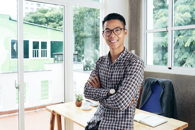 Portrait of smiling confident entrepreneur crossing arms and looking at camera when leaning on desk