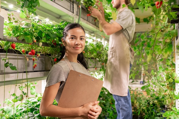 植物の棚に立ってクリップボードを保持している小さな温室の笑顔の自信を持って若い女性労働者の肖像画