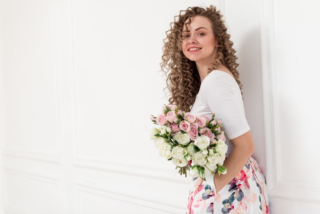 Portrait of smiling curly blonde girl learned on the white wall and holds a bouquet of roses