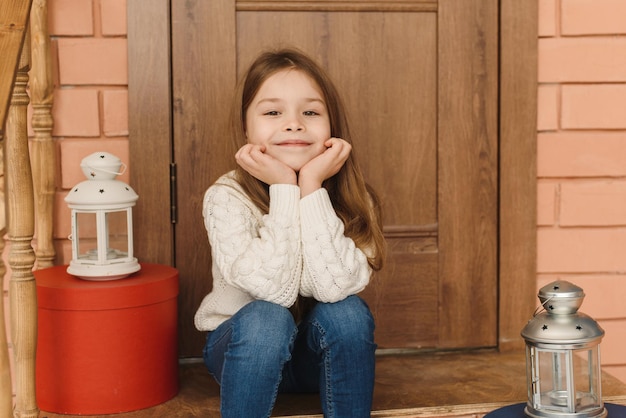 Foto ritratto di una ragazza sorridente e carina seduta sotto il portico di casa con una ghirlanda sulla porta in attesa delle vacanze e delle feste di capodanno