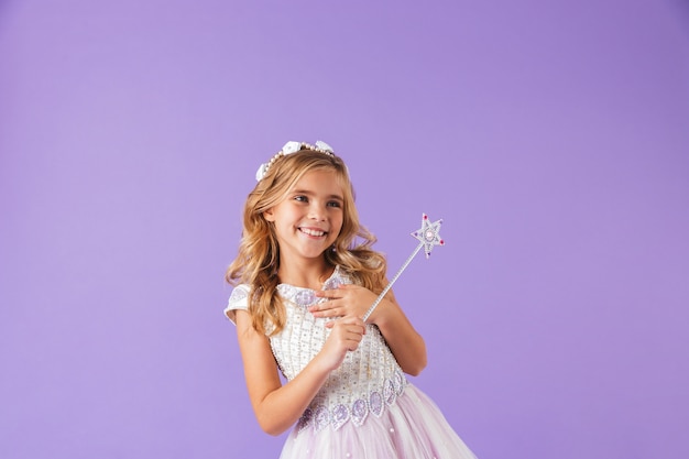 Портрет улыбающейся милой красивой девушки, одетой в платье принцессы, изолированной над фиолетовой стеной, держащей волшебную палочку