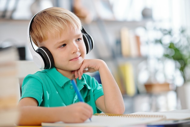 Portrait of smiling cute schoolboy enjoying music in headphones and writing in textbook