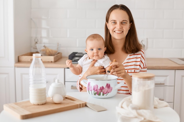 Portrait of smiling dark haired woman sitting at table surrounded with different ingredients for baking satisfied female expressing happiness spending time together with her child