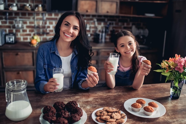 Portrait of smiling daughter and mother holding glasses of milk and cupcakes in the hands looking at the camera on the kitchen