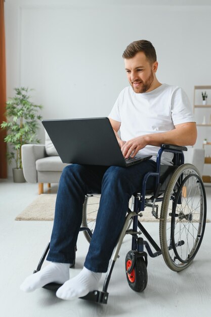 Portrait of smiling disabled male sitting in wheelchair and working on laptop from home Young worker with special needs Freelancer and people with disabilities concept