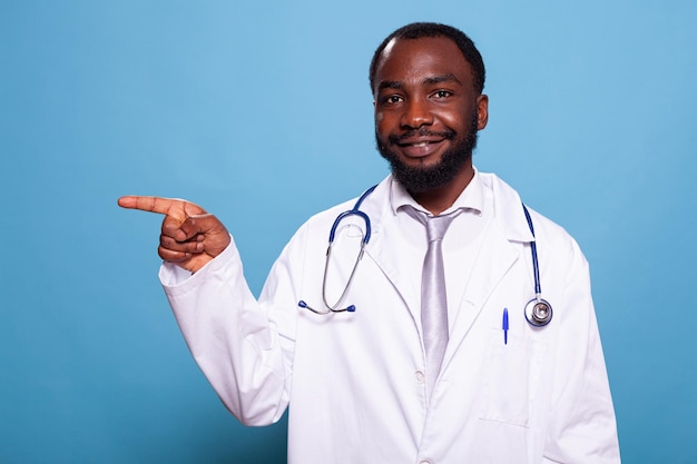 Portrait of smiling doctor wearing hospital uniform and stethoscope pointing next to him on blue background. Smiling medic doing hand gesture with index finger to his right side.