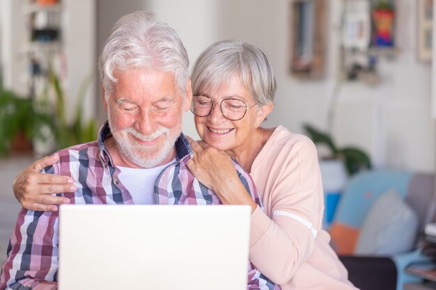 Portrait of smiling elderly couple browsing together on laptop sitting at home Joyful and beautiful senior couple white haired