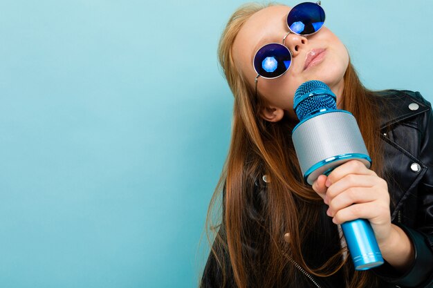 Ritratto di una ragazza europea sorridente in occhiali da sole che canta con un microfono sulla parete blu-chiaro