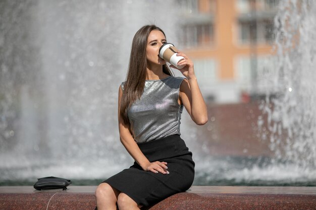 Portrait of smiling european woman in a shiny gray T-shirt strolling through city street with  takeaway coffee in hands