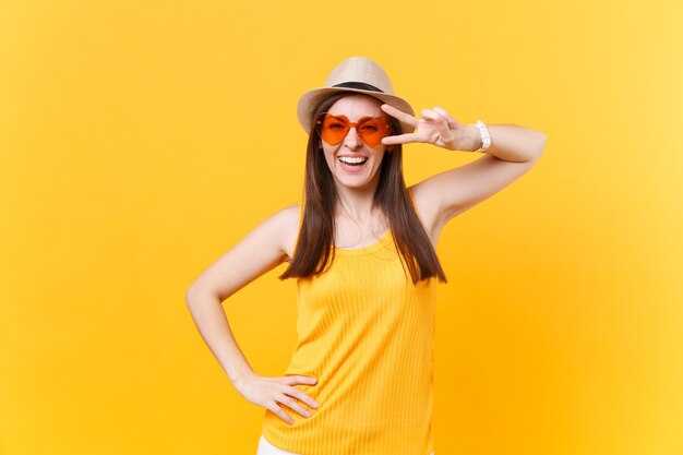 Portrait of smiling excited young woman in straw summer hat, orange glasses showing victory sign, copy space isolated on yellow background. People sincere emotions, lifestyle concept. Advertising area