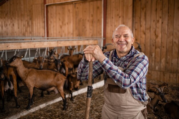 Portrait of smiling farmer or cattleman standing in farmhouse while goat domestic animals eating