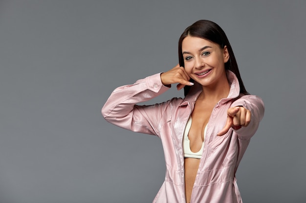 Portrait of a smiling fashion girl in pink overalls pointing her finger at empty copy space. Copy space, gray background. Fashion concept.