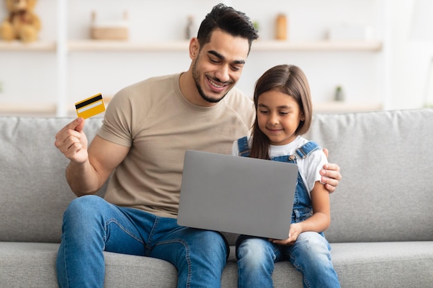 Portrait of smiling father and kid shopping together on Internet, using laptop and credit card, sitting and hugging on the couch at home, looking at computer screen and smiling, order food, copy space