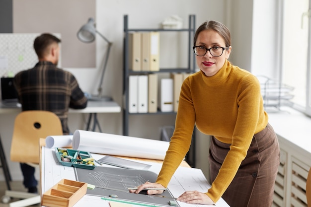 Portrait of smiling female architect drawing blueprints and plans while standing at desk in office and , 