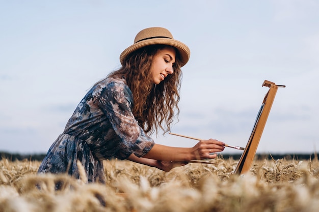 Ritratto dell'artista femminile sorridente con capelli ricci in cappello. la donna disegna un'immagine di un paesaggio in un campo di grano. copia spazio