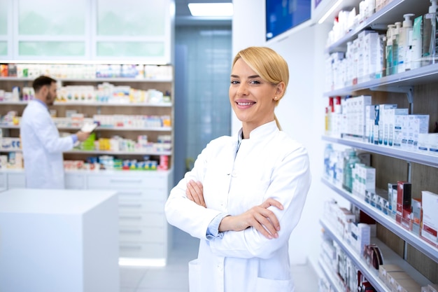 Portrait of smiling female blonde pharmacist standing in pharmacy shop or drugstore with her arms crossed.