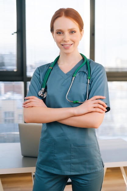 窓の背景に立っている青緑色の制服を着た笑顔の女性医師の肖像画