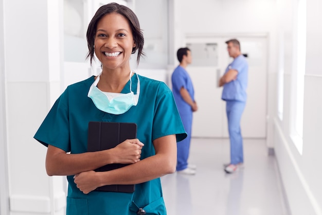 Portrait Of Smiling Female Doctor Wearing Scrubs In Hospital Corridor Holding Digital Tablet