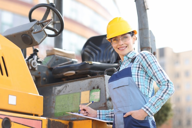 Portrait of smiling female foreman in helmet with documents and pen on background of asphalt paver