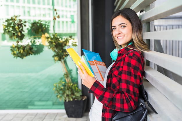 Portrait of a smiling female university student holding books in hand leaning on wall