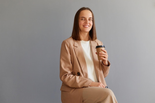 Portrait of smiling female wearing beige jacket posing isolated over gray background sitting holding disposable cup with take away coffee looking at camera with toothy smile