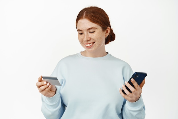 Portrait of smiling female with red hair and freckles, mobile phone and credit card with happy relaxed face on white