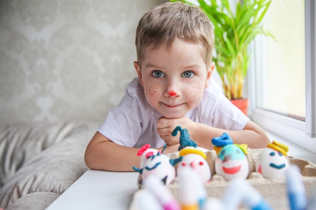 Portrait of a smiling four year old boy with a painted face lying on the windowsill in front of decorated Easter eggs