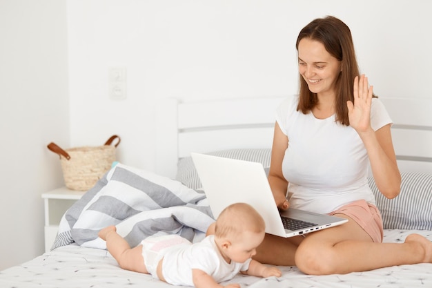 Portrait of smiling friendly woman wearing white t shirt sitting on bed with her toddler and working online, holding notebook, having video call, waving hand, saying hello or bye.