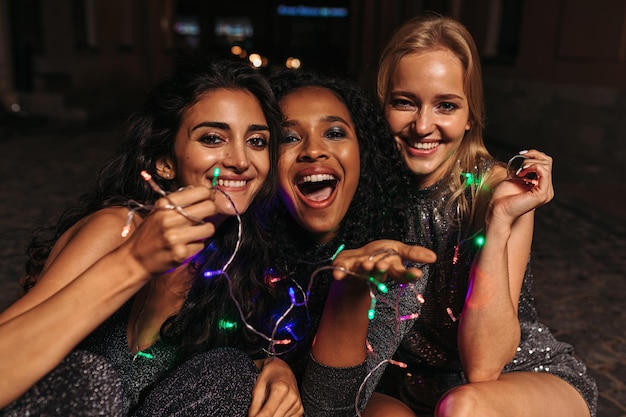 Photo portrait of smiling friends with illuminated lighting equipment sitting in city at night