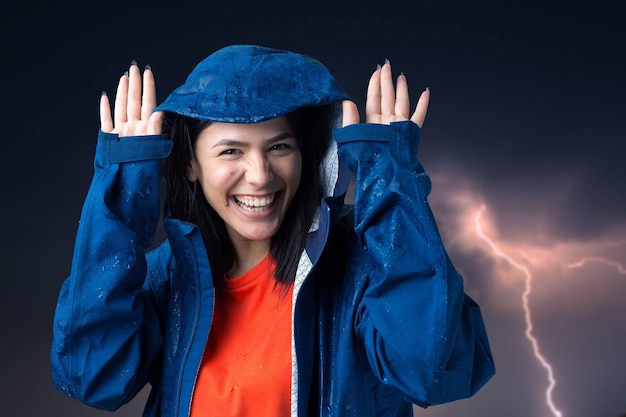 Portrait of a smiling girl dressed in blue raincoat in drops posing with hood against the background of a gloomy sky with lightning