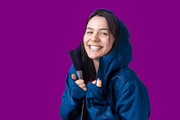 Portrait of a smiling girl dressed in blue raincoat in drops posing with hood on grey background in a studio