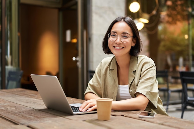 Portrait of smiling girl in glasses sitting with laptop in outdoor cafe drinking coffee and working