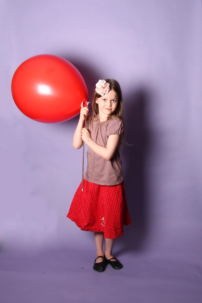 Photo portrait of smiling girl holding red balloon while standing against purple background