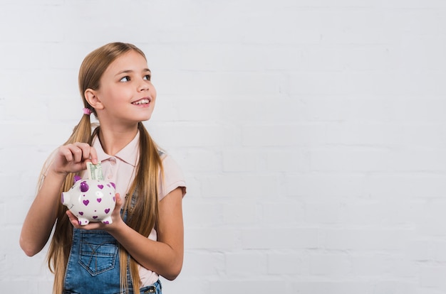 Portrait of a smiling girl inserting currency note in white piggybank looking away