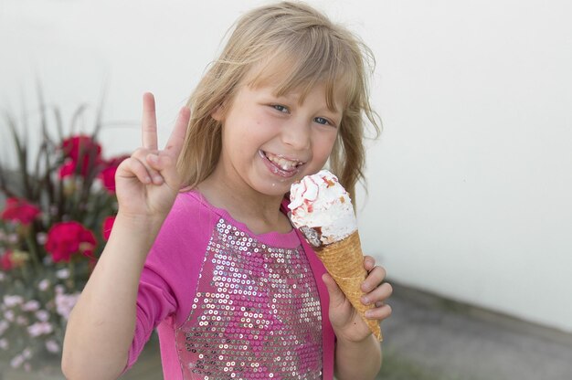 Photo portrait of smiling girl showing peace sign while eating ice cream by wall