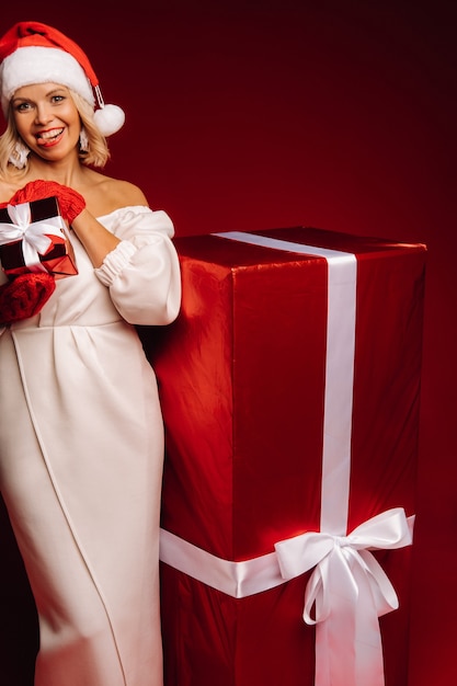 Портрет улыбающейся девушки в белом платье и шляпе Санты с рождественским подарком на красном фоне.