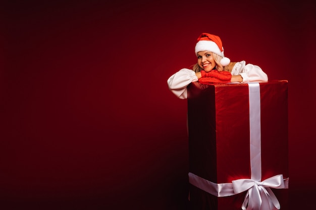 Portrait of a smiling girl in a white dress and Santa hat with a huge Christmas gift on a red background.