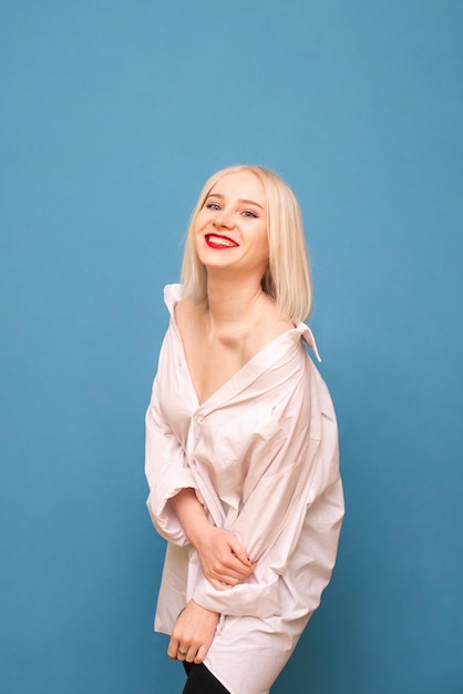 Ritratto di ragazza sorridente in camicia bianca maschile su sfondo blu che posa alla macchina fotografica con la faccia felice