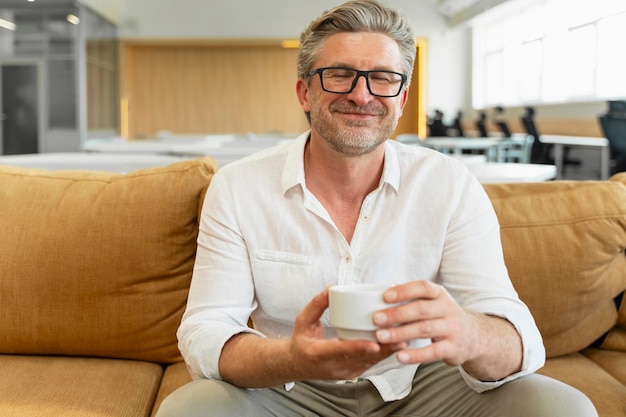 Portrait of smiling gray haired businessman sitting on comfortable sofa drinking coffee