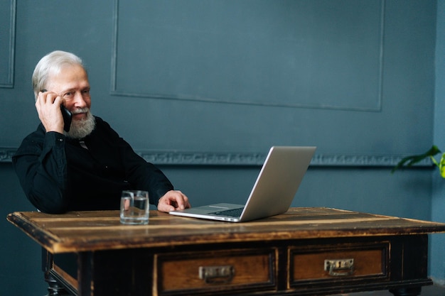 Photo portrait of smiling gray-haired mature older businessman talking on mobile phone sitting at table with laptop computer. bearded cheerful senior aged male having conversation on smartphone .