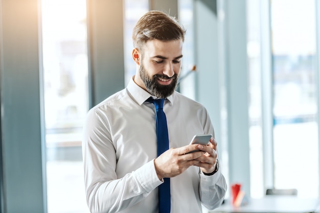 Portrait of smiling handsome bearded businessman in shirt and tie using smart phone while standing in office next to window.