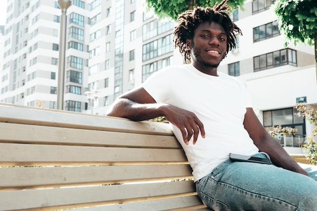 Portrait of smiling handsome hipster modelUnshaven African man dressed in white summer tshirt and jeans Fashion male with dreadlocks hairstyle posing on the street background