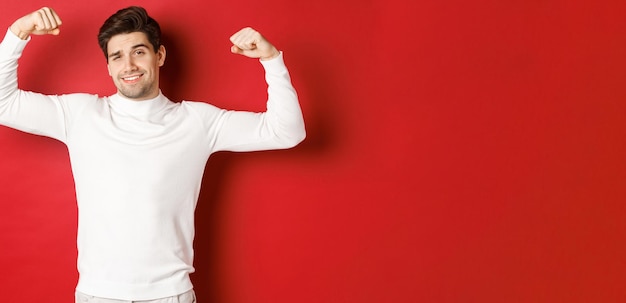 Portrait of smiling handsome man in white sweater, flexing biceps and bragging with strength, show-off strong muscles after workout, standing over red background