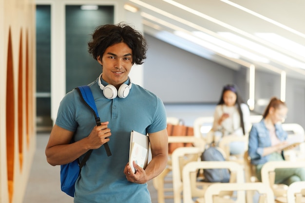 Портрет улыбающегося красивого студента с грязными волосами, стоящего с рюкзаком и ноутбуком в современной университетской комнате