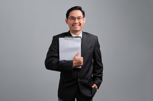 Portrait of smiling handsome young businessman in formal suit and glasses holding clipboard isolated on grey background