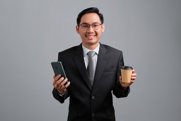 Portrait of smiling handsome young businessman in formal suit and glasses holding mobile phone and paper coffee cup isolated on grey background