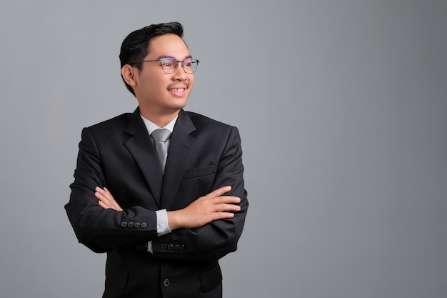 Portrait of smiling handsome young businessman in formal suit and glasses standing with crossed arms and looking away isolated on grey background