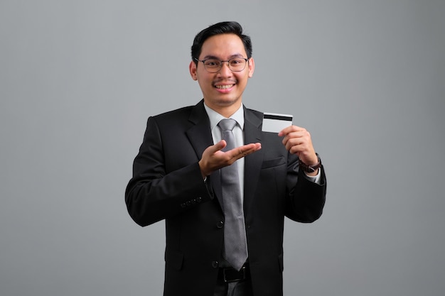 Portrait of smiling handsome young businessman in formal suit showing credit card isolated on grey background